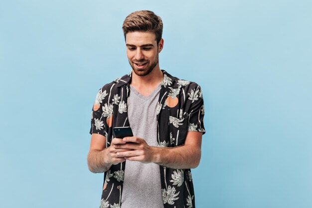 Giovane con acconciatura alla moda e barba in maglietta grigia fresca e camicia a maniche corte moderna estiva che esamina il telefono e sorride