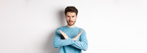 Giovane con la barba che sembra serio facendo un gesto incrociato per fermare qualcosa proibire l'azione in piedi