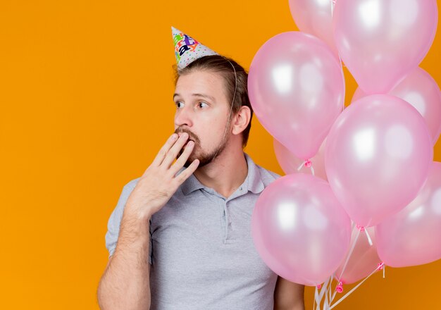 Giovane con tappo di festa che celebra la festa di compleanno che tiene mazzo di palloncini che osserva da parte che copre la bocca con la mano sorpreso e stupito sull'arancia