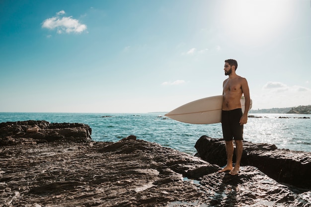 Giovane con tavola da surf sulla pietra vicino mare