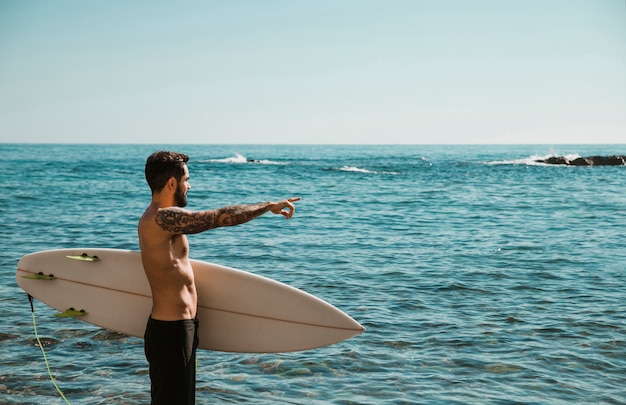 Giovane con tavola da surf sulla spiaggia che punta all&#39;acqua