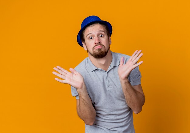 Giovane con un cappello che celebra la festa di compleanno che sembra confuso allargando le braccia ai lati sopra l'arancio