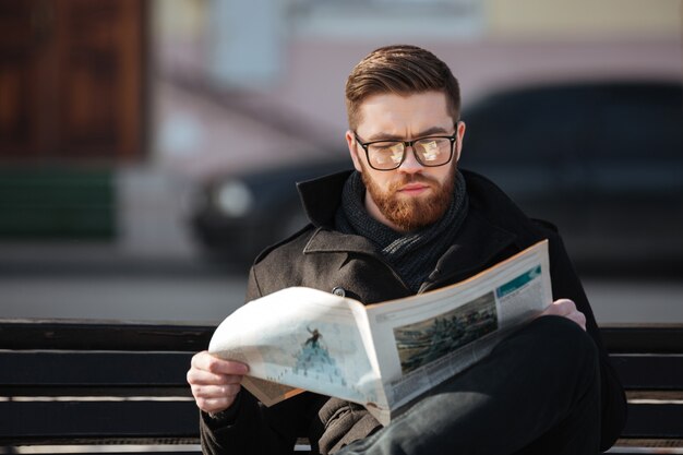 Giovane concentrato che si siede sul banco e che legge giornale all'aperto