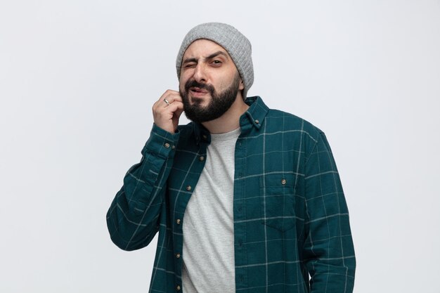 Giovane confuso che indossa un cappello invernale guardando la fotocamera che graffia il viso con un occhio chiuso isolato su sfondo bianco