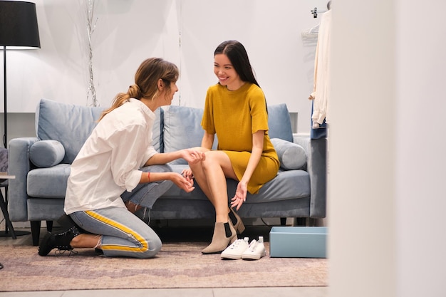 Giovane consulente di moda che aiuta gioiosamente a provare gli stivali per una ragazza asiatica allegra in uno showroom moderno