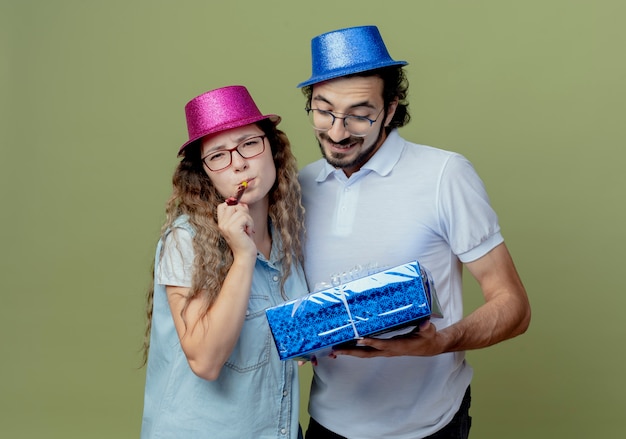 Giovane coppia che indossa rosa e blu cappello ragazza che soffia fischio e ragazzo che tiene e guardando la confezione regalo isolata sulla parete verde oliva