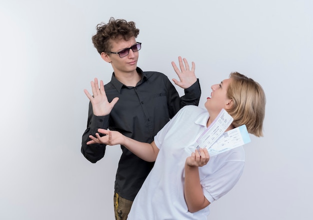 Giovane coppia di turisti uomo confuso e scontento guardando la sua ragazza con i biglietti aerei nelle mani in piedi sopra il muro bianco