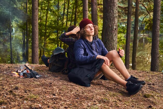 Giovane coppia hipster che fa un'escursione nella foresta e si siede schiena contro schiena mentre si riscalda vicino a un falò al campo. Concetto di viaggio, turismo ed escursione.