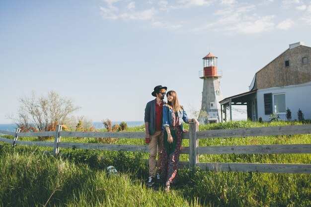 Giovane coppia hipster stile indie innamorato che cammina in campagna, tenendosi per mano, faro sullo sfondo, calda giornata estiva, soleggiato, vestito bohémien, cappello