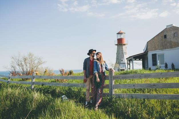 Giovane coppia hipster stile indie innamorato che cammina in campagna, tenendosi per mano, faro sullo sfondo, calda giornata estiva, soleggiato, vestito bohémien, cappello