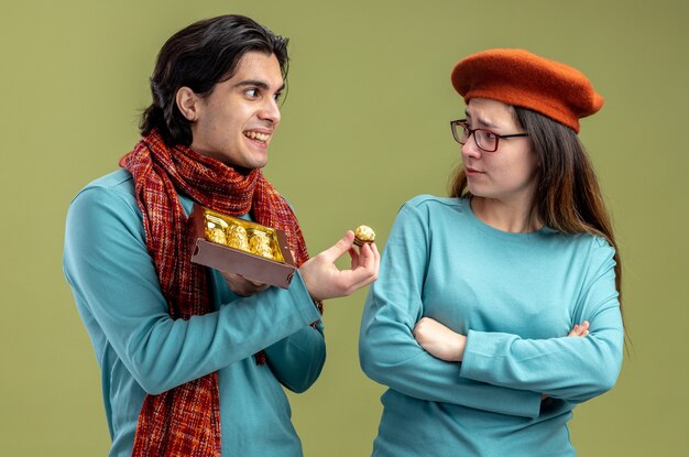 Giovane coppia il giorno di san valentino ragazzo che indossa una sciarpa ragazza che indossa un cappello ragazzo sorridente che dà scatola di caramelle isolato su sfondo verde oliva
