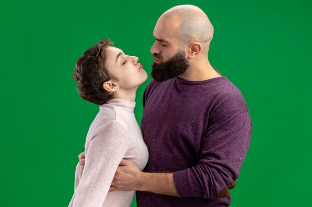 giovane coppia in abiti casual donna con i capelli corti e l'uomo barbuto felice innamorato insieme abbracciando andando a baciare celebrando il giorno di San Valentino in piedi su sfondo verde