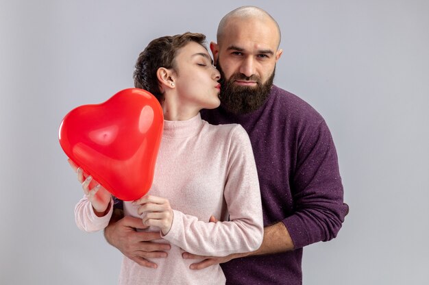 giovane coppia in abiti casual uomo e donna azienda palloncino a forma di cuore donna che bacia il suo ragazzo felice che celebra il giorno di San Valentino in piedi sopra il muro bianco