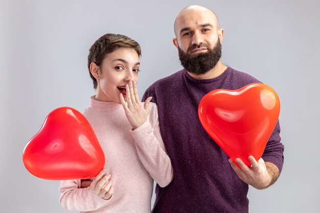 giovane coppia in abiti casual uomo e donna in possesso di palloncini a forma di cuore guardando la fotocamera felice e sorpreso per celebrare il giorno di San Valentino in piedi sul muro bianco