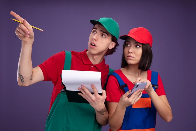 Giovane coppia in uniforme da operaio edile e berretto impressionato ragazzo che tiene matita e appunti puntando verso l'alto ragazza seria che tiene il telefono cellulare entrambi alzando lo sguardo isolato sul muro viola