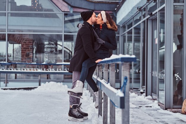 Giovane coppia innamorata, appuntamento alla pista di pattinaggio, una ragazza seduta su un guardrail e bacio con il suo ragazzo.