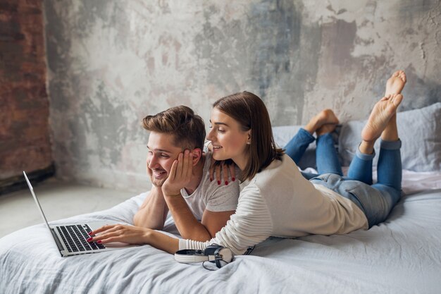 Giovane coppia sorridente sdraiato sul letto a casa in abbigliamento casual, guardando in laptop, uomo e donna che trascorrono del tempo felice insieme, rilassante
