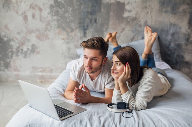 Giovane coppia sorridente sdraiato sul letto a casa in abito casual, guardando in laptop, uomo e donna che trascorrono del tempo felice insieme, rilassante
