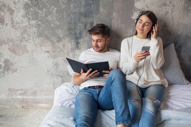 Giovane coppia sorridente seduto sul letto a casa in abbigliamento casual leggendo il libro che indossa jeans, uomo che legge il libro, donna che ascolta la musica in cuffia, trascorrere del tempo romantico insieme