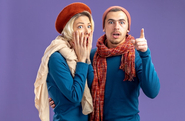 Giovane coppia spaventata che indossa un cappello con sciarpa il giorno di san valentino ragazza ha coperto la bocca con un ragazzo a mano che punta alla telecamera isolata su sfondo blu