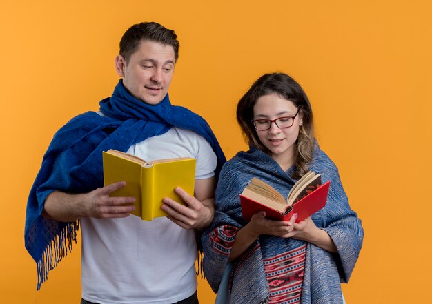 Giovane coppia uomo e donna con coperte in possesso di libri sorridente felice e positivo in piedi sopra la parete arancione
