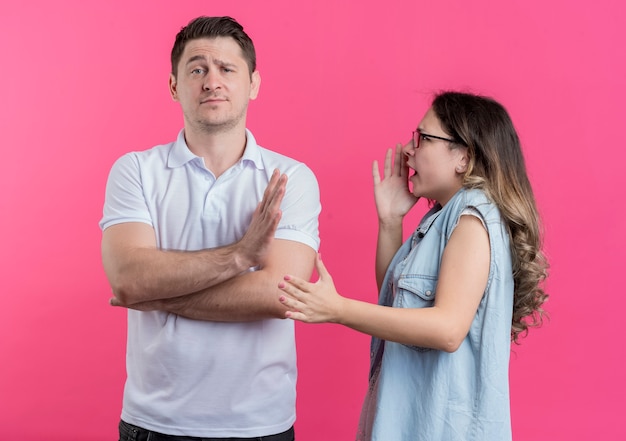 Giovane coppia uomo e donna in abiti casual donna dispiaciuta che grida al suo fidanzato mentre fa il gesto di arresto in piedi sopra il muro rosa