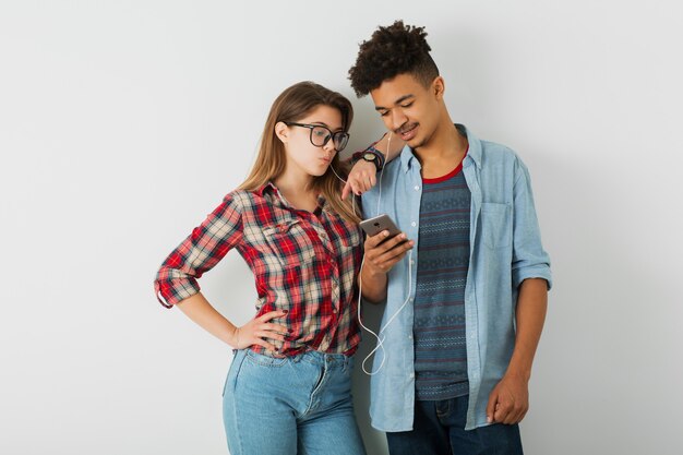 Giovane coppia, uomo nero hipster, ragazza caucasica su sfondo bianco isolato, vestito elegante, acconciatura afro, sorridente, amici insieme divertendosi, ascoltando musica sugli auricolari, tenendo il telefono intelligente