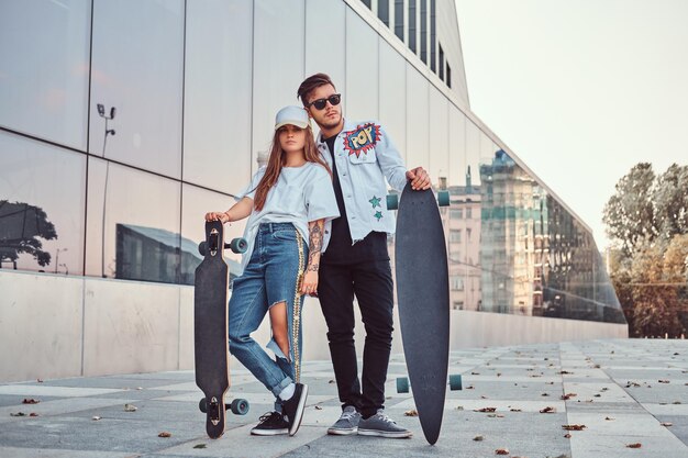 Giovane coppia vestita con abiti alla moda in posa con skateboard vicino al grattacielo.