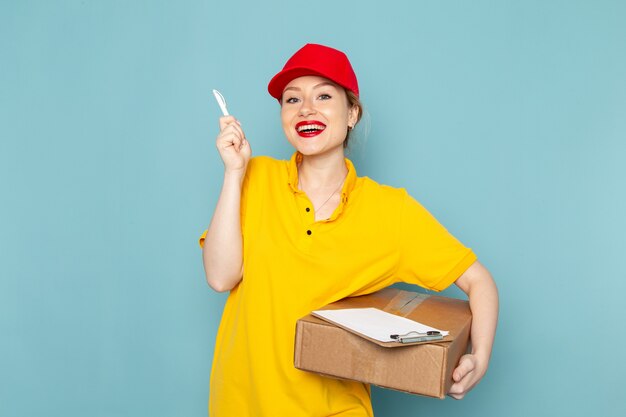 Giovane corriere femminile di vista frontale in camicia gialla e blocco note rosso del pacchetto della tenuta del mantello che sorride sul lavoro di lavoro dello spazio blu