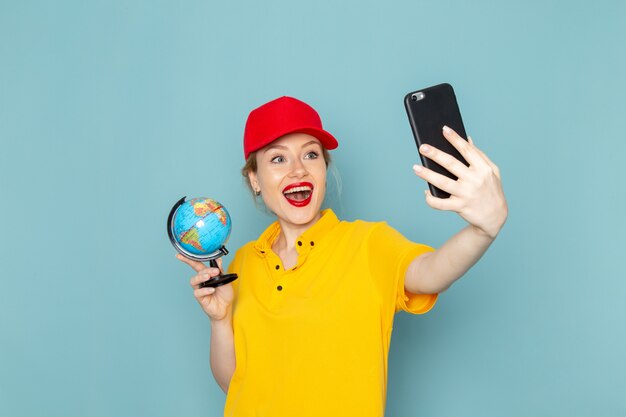 Giovane corriere femminile di vista frontale in camicia gialla e mantello rosso che prende un selfie con il globo che sorride sullo spazio blu