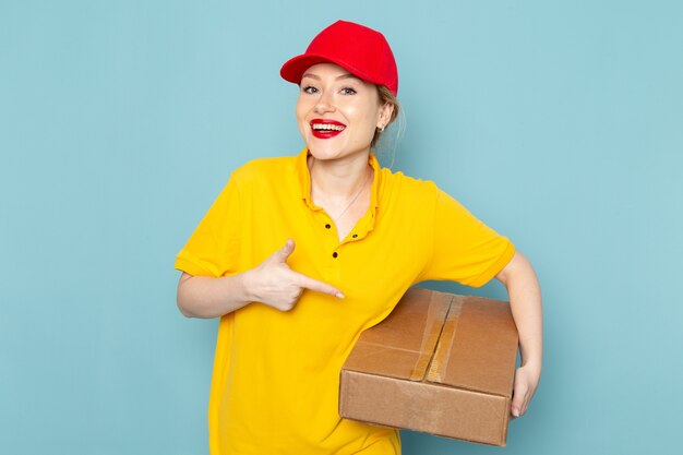 Giovane corriere femminile di vista frontale in camicia gialla e mantello rosso che sorride e che tiene pacchetto sul lavoro dello spazio blu