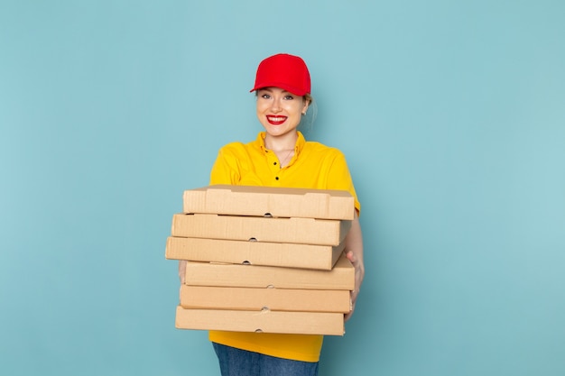 Giovane corriere femminile di vista frontale in camicia gialla e mantello rosso che tiene i pacchetti che sorridono sul lavoro dello spazio blu