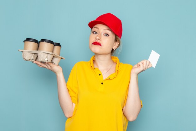 Giovane corriere femminile di vista frontale in camicia gialla e mantello rosso che tiene la carta bianca delle tazze di caffè di plastica sul lavoro di lavoro dello spazio blu