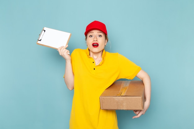 Giovane corriere femminile di vista frontale in camicia gialla e pacchetto rosso della tenuta del mantello e blocco note sul lavoratore di lavoro dello spazio blu