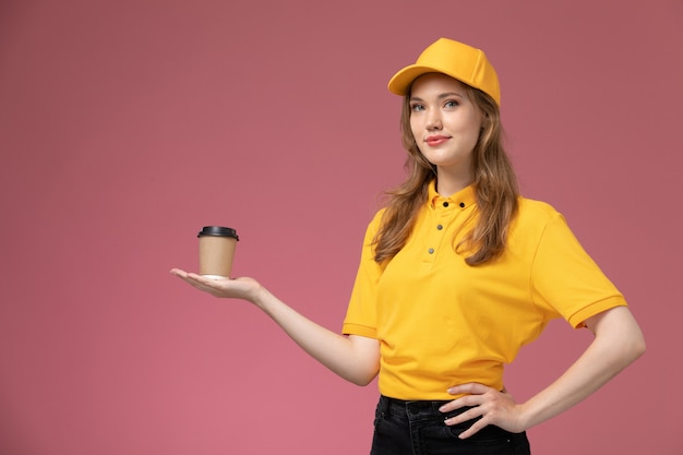 Giovane corriere femminile di vista frontale in capo giallo uniforme giallo che tiene caffè con il sorriso sulla lavoratrice di servizio di consegna uniforme sfondo rosa scuro