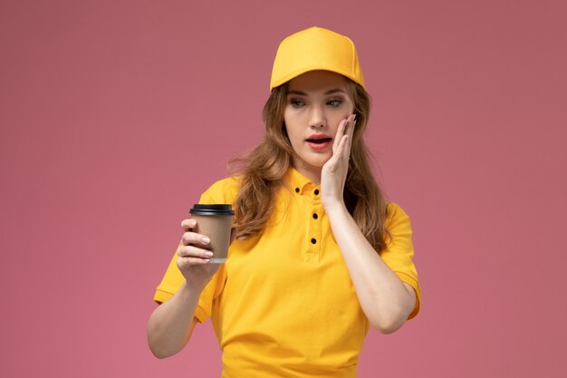 Giovane corriere femminile di vista frontale in capo giallo uniforme giallo che tiene caffè sul colore di servizio di consegna uniforme sfondo rosa scuro