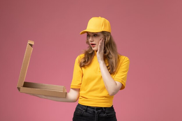 Giovane corriere femminile di vista frontale in mantello giallo uniforme giallo che tiene la scatola di consegna del cibo aprendolo sul colore di servizio di lavoro di consegna uniforme di sfondo rosa scuro