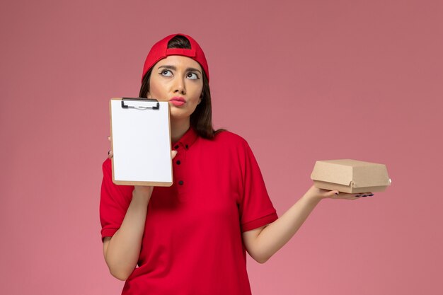 Giovane corriere femminile di vista frontale in mantello rosso uniforme con poco pacchetto di cibo di consegna e blocco note sulle sue mani pensando sulla parete rosa