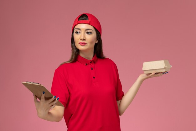 Giovane corriere femminile di vista frontale in mantello rosso uniforme con poco pacchetto di cibo di consegna e blocco note sulle sue mani sulla parete rosa