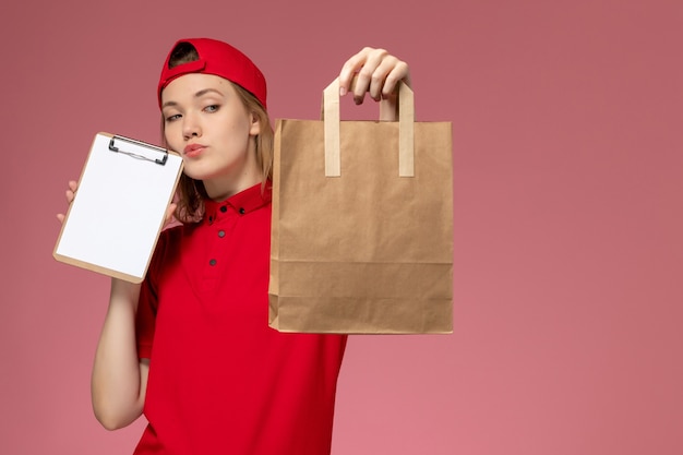 Giovane corriere femminile di vista frontale in pacchetto di cibo di consegna uniforme rosso della tenuta e blocco note che pensa sulla parete rosa-chiaro