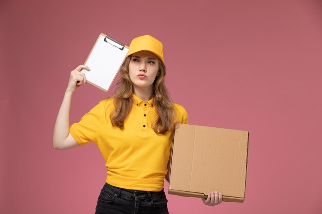 Giovane corriere femminile di vista frontale in scatola dell'alimento della tenuta dell'uniforme gialla e blocco note che pensa sull'operaio di servizio di consegna dell'uniforme di lavoro dello scrittorio rosa scuro