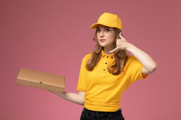 Giovane corriere femminile di vista frontale in scatola dell'alimento di consegna della tenuta della tenuta uniforme gialla che posa sull'operaio di servizio di consegna dell'uniforme di lavoro dello scrittorio rosa