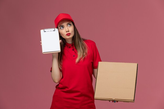 Giovane corriere femminile di vista frontale in scatola di cibo di consegna della tenuta uniforme rossa con il blocco note che pensa sulla società uniforme di servizio di consegna del fondo rosa