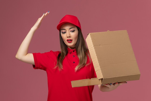 Giovane corriere femminile di vista frontale in scatola di cibo di consegna della tenuta uniforme rossa sulla società uniforme di lavoro di consegna del servizio di scrivania rosa chiaro