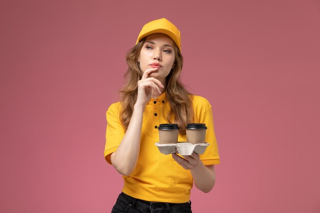 Giovane corriere femminile di vista frontale in tazze di caffè della tenuta dell'uniforme gialla che posano con l'espressione di pensiero sul lavoratore di servizio di lavoro di consegna dell'uniforme dello scrittorio rosa scuro