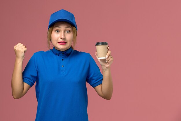 Giovane corriere femminile di vista frontale in uniforme blu che posa che tiene la tazza di caffè marrone di consegna, donna di consegna dell'uniforme di lavoro di servizio