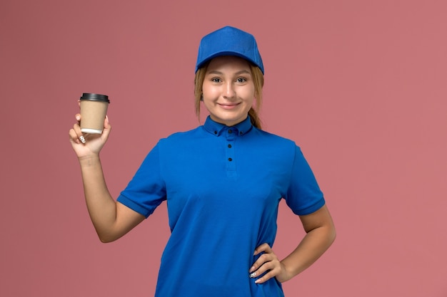Giovane corriere femminile di vista frontale in uniforme blu che posa la tazza di caffè di consegna della tenuta sulla donna di consegna dell'uniforme di lavoro di servizio della parete rosa