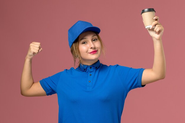 Giovane corriere femminile di vista frontale in uniforme blu che posa tenendo la tazza di caffè che flette, colore dell'operaio di lavoro della donna di consegna dell'uniforme di servizio