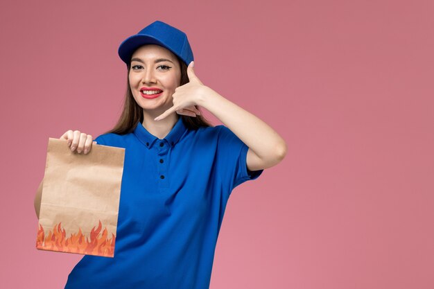 Giovane corriere femminile di vista frontale in uniforme blu e mantello che tiene il pacchetto alimentare di carta con il sorriso sul pavimento rosa