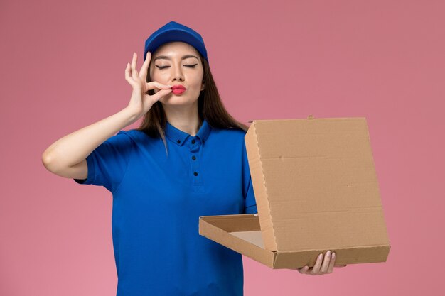 Giovane corriere femminile di vista frontale in uniforme blu e mantello che tiene la scatola vuota di consegna del cibo sulla parete rosa